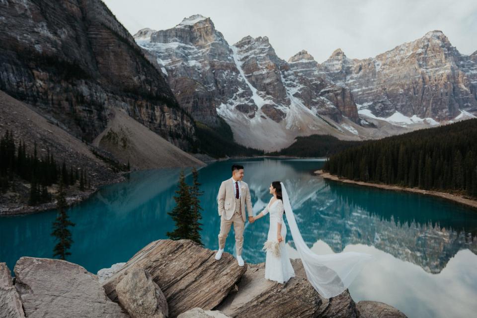 A bride and groom look at each other and hold hands on a rock in front of a pond and mountains.