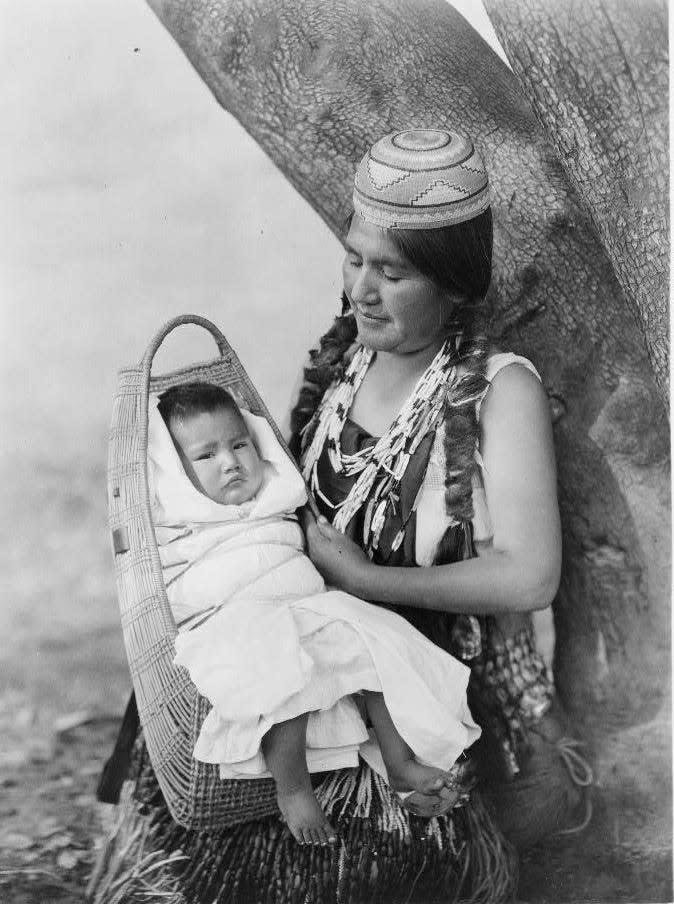 A Native Hupa woman holds a baby on her lap in a traditional carrier.