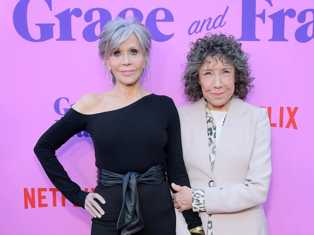 Jane with short grey hair in a black blouse and Lily with curly grey hair in a cream blazer both smiling in front of a purple "Grace and Frankie" backdrop.