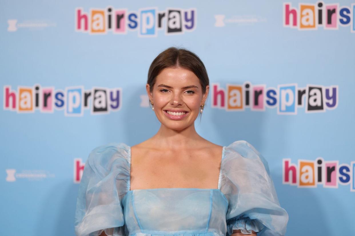 olivia frazer, a woman with brown hair back in a bun smiles at the camera, wearing a blue dress with puff sleeves