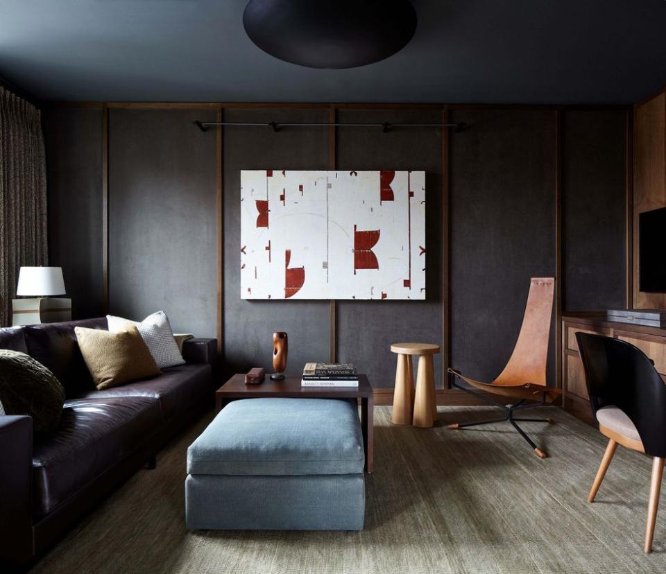 minimalist living space with dark brown leather sofa with a oblong velvet pouf beneath a small table in front a light washed stool and modern chairs a large white and red canvas sits in the center of the wall opposite