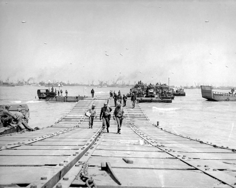 People in uniforms walk across a series of boxes fastened together and floating on the water. Ships are visible in the background.