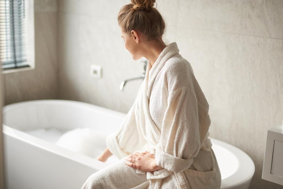 person wearing white bathrobe sitting on edge of tub filled with bubbles