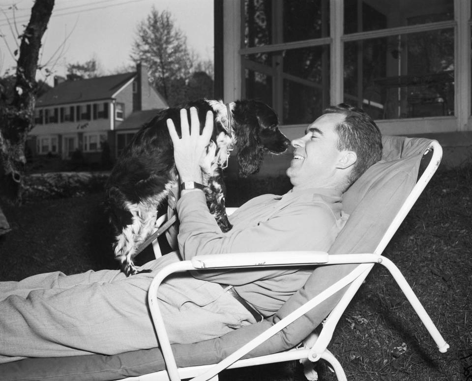 A man lies in a reclining chair and holds a dog up to his face in a black and white photo.