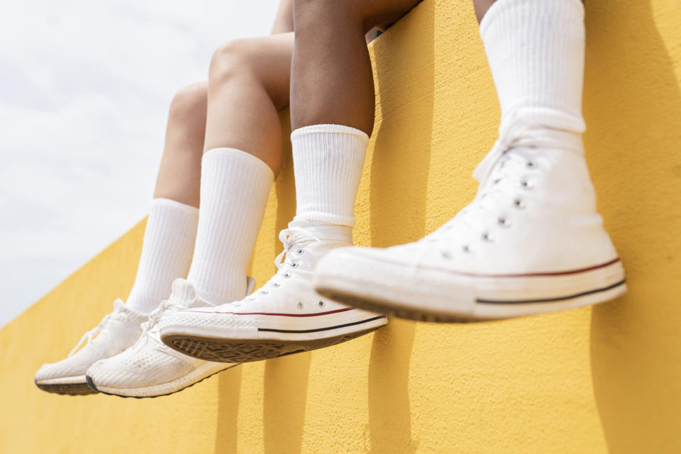 Two people in white socks and white sneakers sit on a yellow wall, their legs dangling over the edge