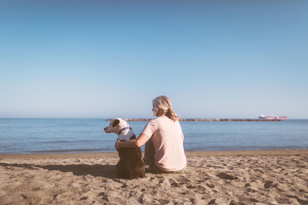 Senior woman sitting on sand and enjoying sunny day at the beach with pet dog