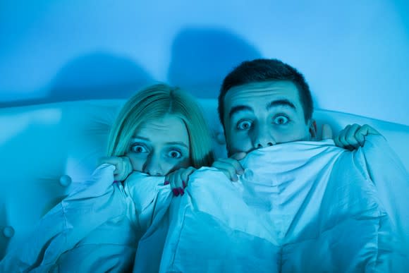 Couple hiding under a blanket with scared expressions.