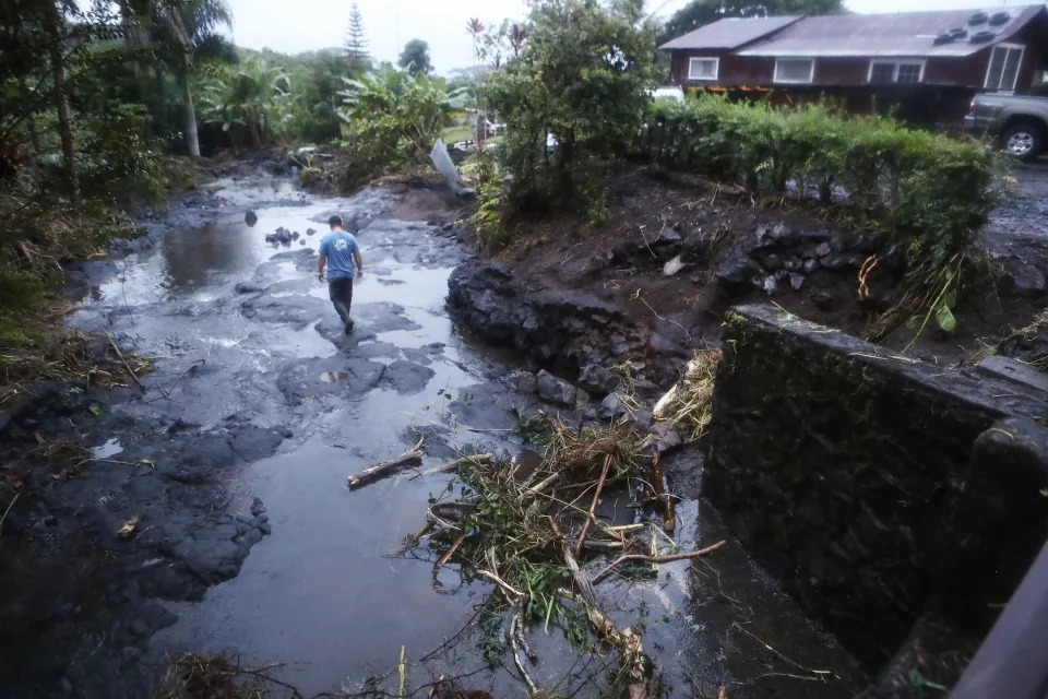 A man picks his way through puddles on a road next to a home damaged by flooding.