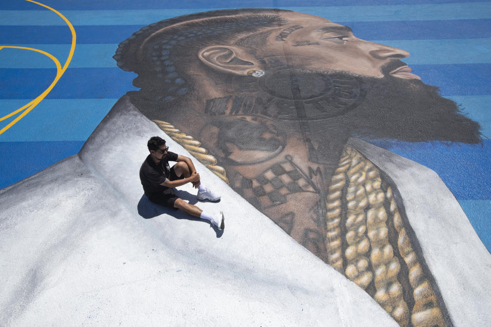 FILE - In this April 17, 2019 file photo, mural artist Gustavo Zermeno Jr. poses for photos on a basketball court mural he dedicated to slain rapper Nipsey Hussle in Los Angeles. Hussle, 33, was shot and killed outside his Los Angeles clothing store on March 31, 2019. A year after Hussle's death, his popularity and influence are as strong as ever. He won two posthumous Grammys in January, he remains a favorite of his hip-hop peers and his death has reshaped his hometown of Los Angeles in some unexpected ways. (AP Photo/Jae C. Hong, File)