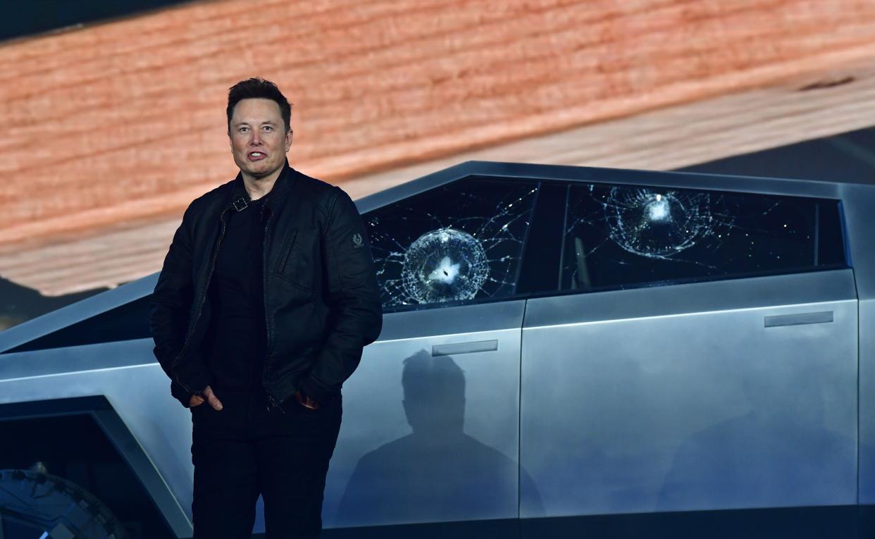 Elon Musk standing in front of a Cybertruck with shattered windows.