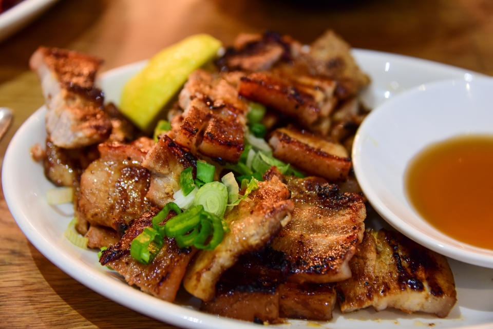 Grilled pork belly pieces garnished with chopped green onions served on a white plate with a side dish of dipping sauce