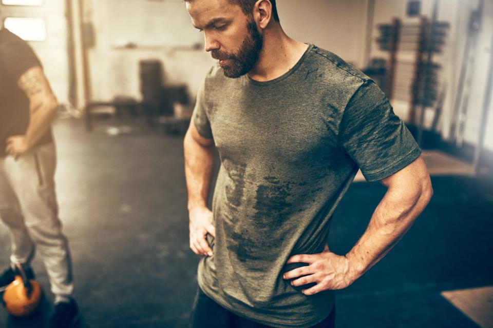 A man in a sweat-soaked shirt and gym shorts with his hands on his hips, looking tired after a workout 