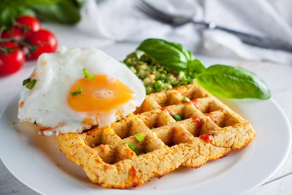 A potato waffle topped with a sunny side up egg next to avocado and fresh basil