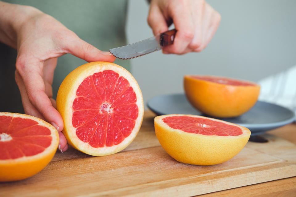 A girl or woman is holding a knife, cutting a red ripe grapefruit in half, on a cutting board, against the background of a wooden kitchen table. The concept of vegetarian, vegan and raw food.