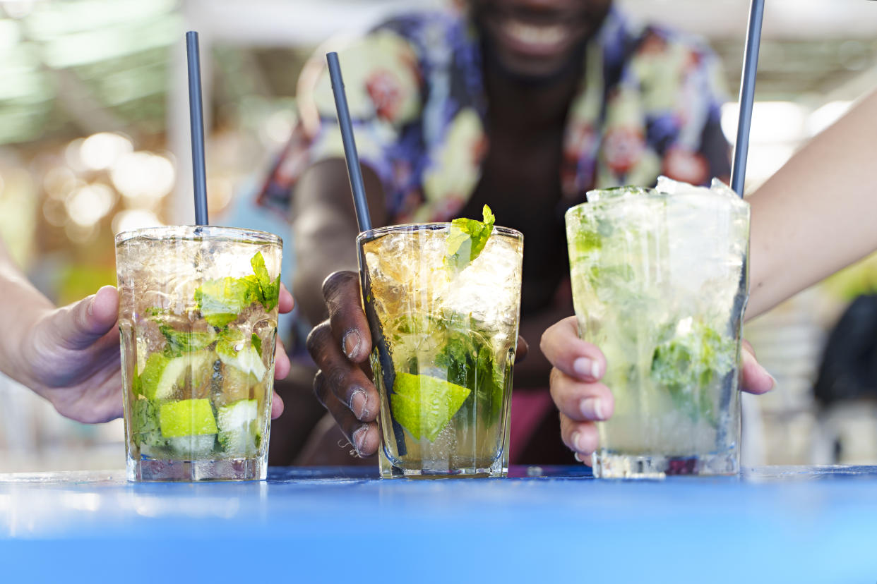 Best friends toasting cocktails. Multiethnic young people drinking long drinks at beach bar. Shallow depth of field with focus on hands holding iced mojitos. Close up shot, focus on the center glass.