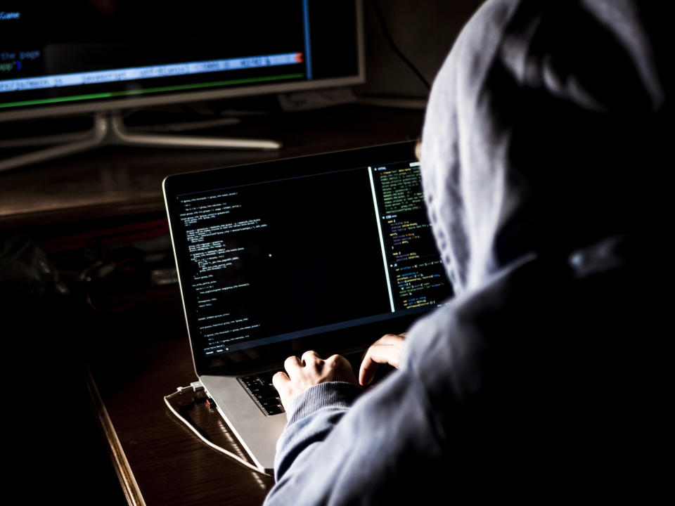 hacker in hoodie typing code in program using laptop in dark studio