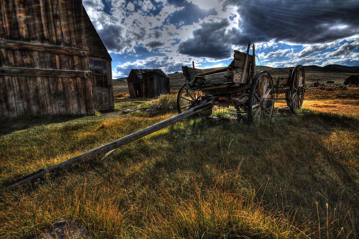 An old wooden wagon in the ghost town of Bodie, California, two buildings on the left, dramatic feel of image, mountains in the background with a blue sky and clouds, sunlight coming through