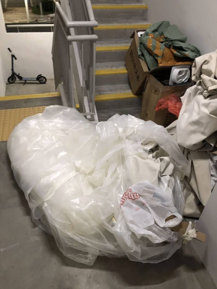 Large plastic bag filled with waste beside stairs, with boxes and a scooter in the background