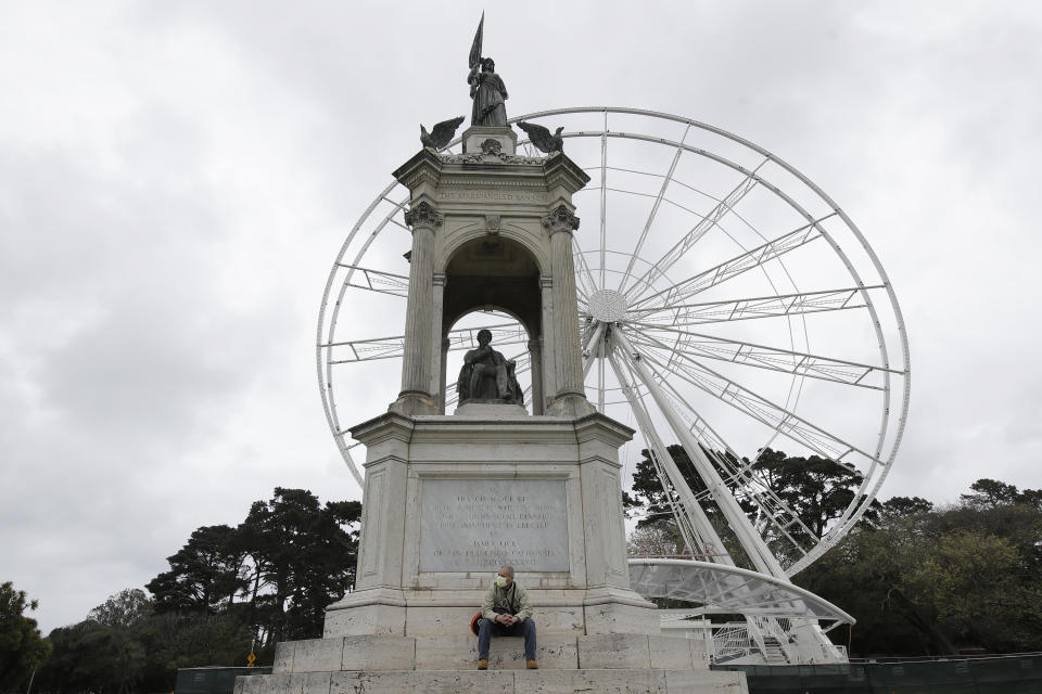 FILE - In this Saturday, March 28, 2020, file photo, Eduardo Vernier wears a mask while sitting under the Francis Scott Key monument at Golden Gate Park in San Francisco, amid the coronavirus outbreak. At rear is the SkyStar Observation Wheel. Golden Gate Park turned 150 on Saturday, April 4. To mark this historic occasion, a virtual concert series is being launched that will bring some of the past outdoor concerts and performances held in the park to people everywhere to enjoy for free. (AP Photo/Jeff Chiu, File)