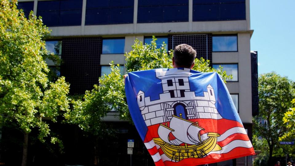 A fan draped in the Serbian flag looks up at the Park Hotel in Melbourne where Novak Djokovic has reportedly been detained