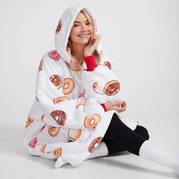 A blonde model wears a white The Oodie with images of iced donuts scattered over it. She has black leggings and white socks