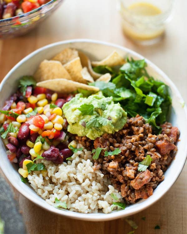 A bowl filled with equal parts rice, lentil walnut taco meat, a corn salad, tortilla chips, and guacamole.