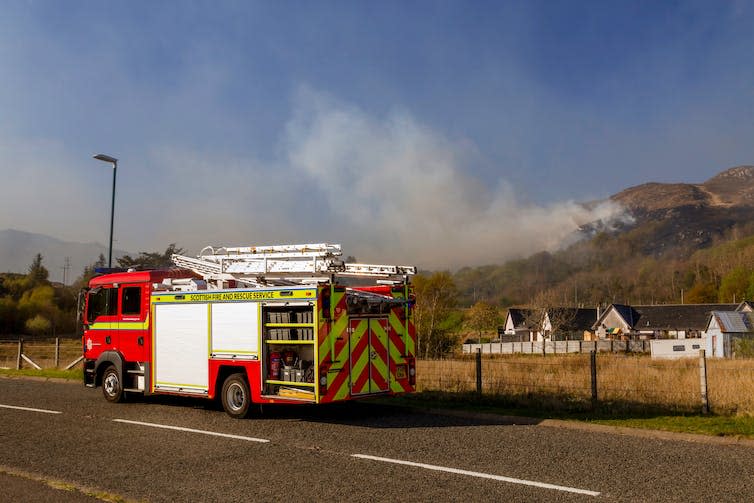 A fire truck parked on a rural road with a grass fire on a hillside in the distance.