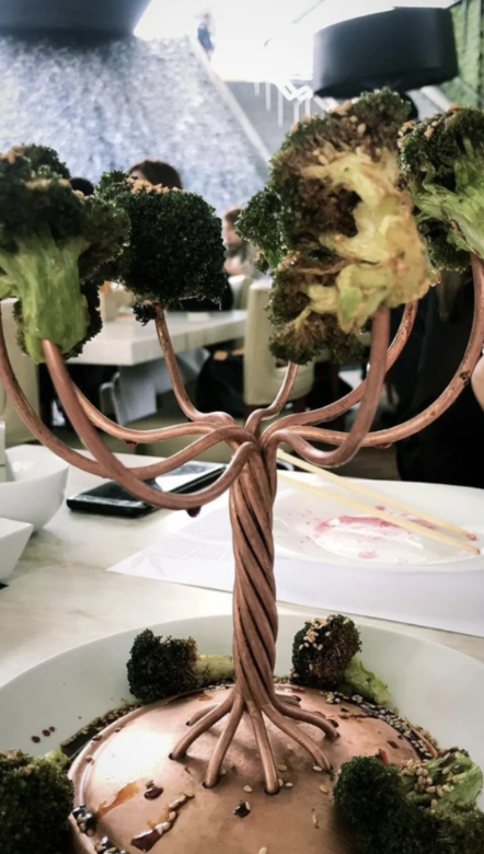 A whimsical metal tree sculpture holding roasted broccoli florets as branches sits on a table in a modern dining setting