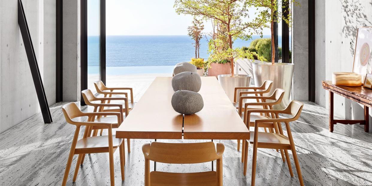 clean spare dining room with a view of a long light wood dining table and chairs with rock like sculptures on it and gray streaked floor with black and white and globe suspected lighting above the table all opening out from floor to ceiling glass doors to a glorious view of the water