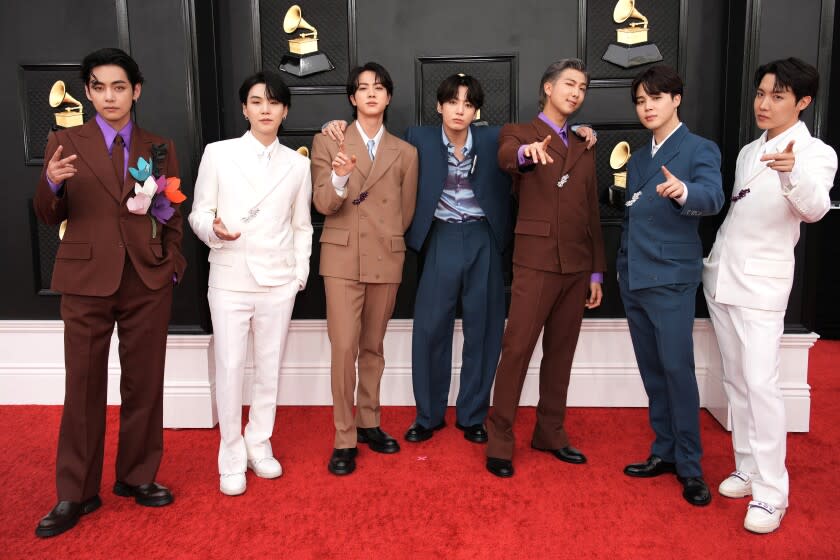 Seven men posing on a red carpet in alternating brown, white and blue suits