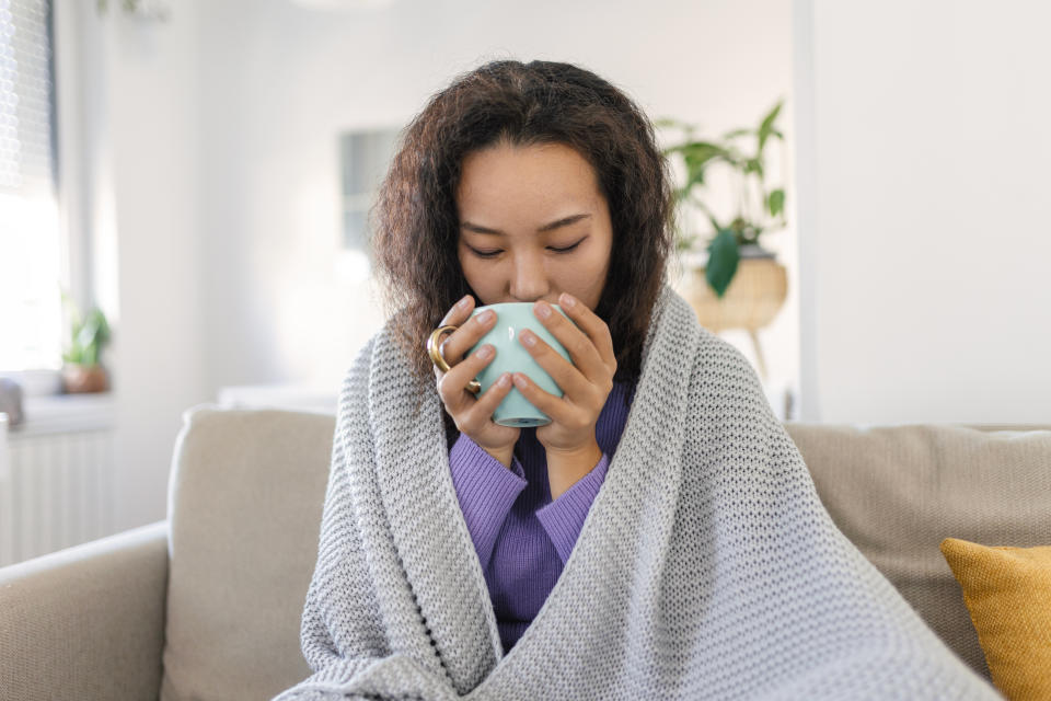 Sick young Asian woman sitting at home in bed with hot cup of tea and handkerchief. Seasonal colds, cough, runny nose, viral infections, home treatment