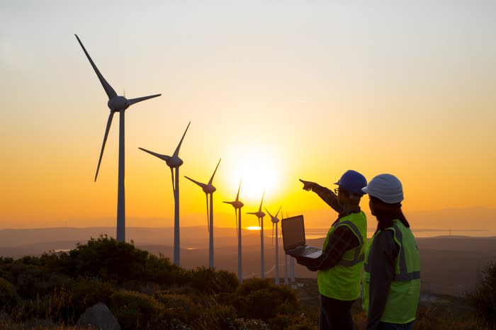 Two people wearing hard hats, with the man holding a laptop and pointing at a row of wind turbines during sunset.