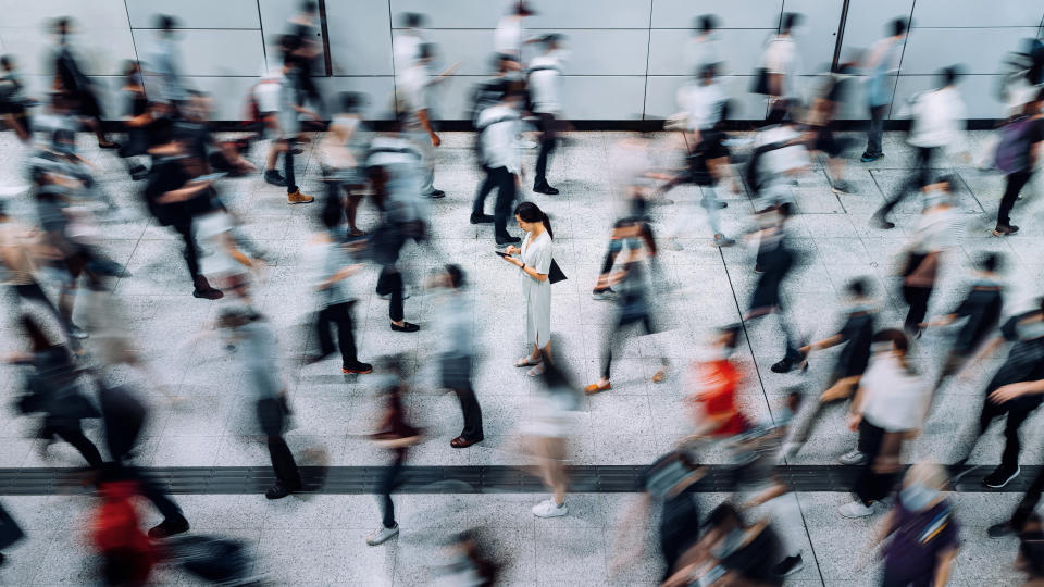 Young Asian woman using smartphone surrounded by commuters rushing by in subway station during office peak hours in the city