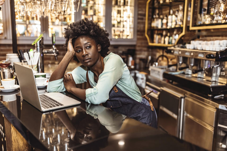 Portrait of upset waitress. She looking aside and sitting behind table while touching her head by palm of hand