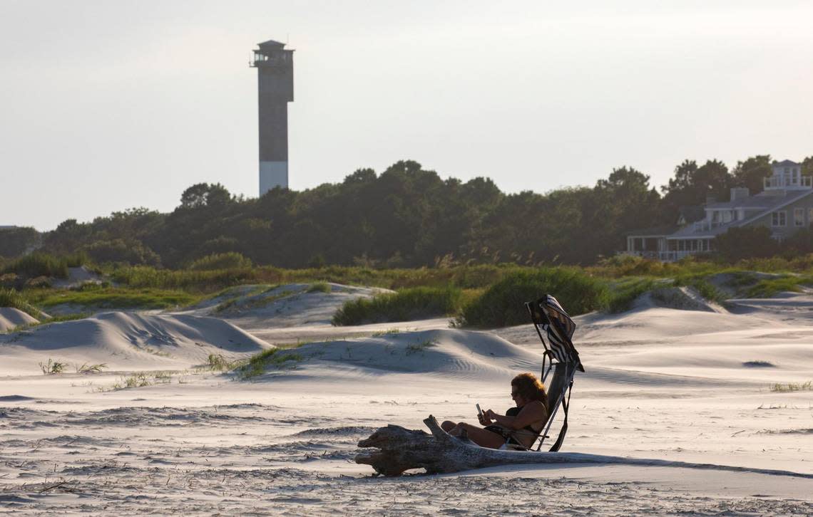 I woman sits on the broad expanse of beach and dunes at Sullivan’s Island with the lighthouse in the background. Shifting sands are having opposite effects on some Charleston area beaches with erosion claiming the Folly Beach shoreline while Sullivan Island’s beach is growing. July 21, 2022.