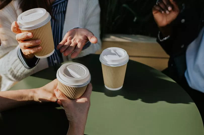 A group of friends / coworkers enjoy hot drinks from takeaway cups at an outdoor cafe / setting.