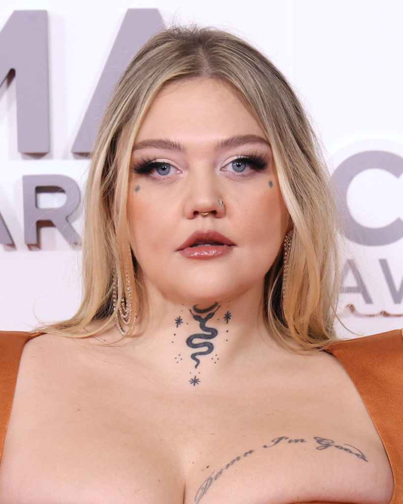 A person with tattoos, long earrings, and makeup poses at a media event, wearing a gown with structured shoulders