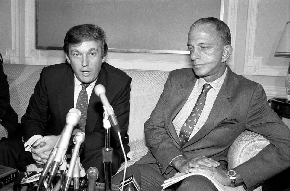 donald trump speaking into a cluster of microphones and roy cohn sits to his left and looks toward him