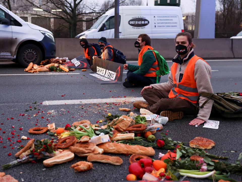 protesters wearing orange vests and face masks sit on a road surrounded by scattered food bread products and produce