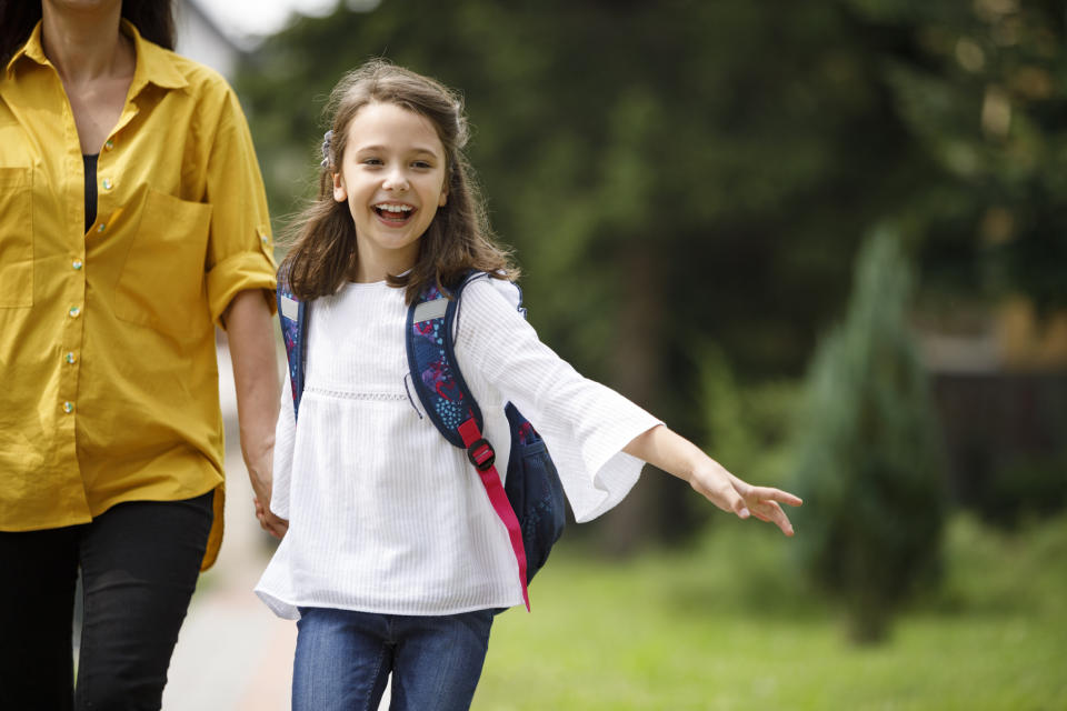 A young girl with a backpack, smiling, walks hand-in-hand with an adult in a casual yellow shirt. They appear to be heading to or from school