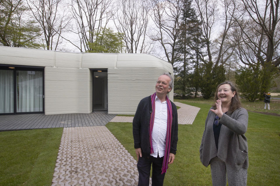 Tenants Elize Lutz, right, and Harrie Dekkers' new home, rear, is a 94-square meters (1,011-square feet) two-bedroom bungalow resembling a boulder with windows in Eindhoven, Netherlands, Friday, April 30, 2021. The fluid, curving lines of its gray walls look natural. But they are actually at the cutting edge of housing construction in the Netherlands and around the world. They were 3D printed at a nearby factory. (AP Photo/Peter Dejong)