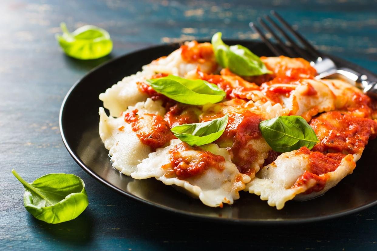 Many ravioli with ripe tomatoes on a round black porcelain plate, a fork next to it, two basil leaves on a rustic ocean blue wooden table