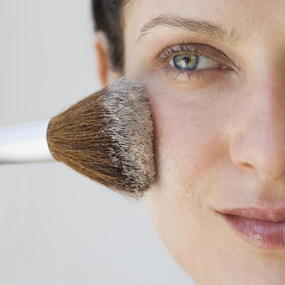 Close-up of a woman's face receiving makeup application with a brush. The image highlights her eye and part of her cheek