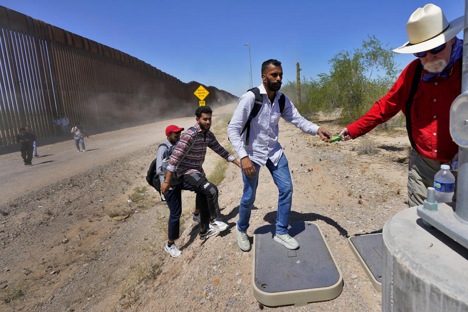 Retired schoolteacher Tom Wingo of Samaritans Without Borders, right, gives snacks and bottles of waters to a group of migrants claiming to be from India, who just crossed the border wall, Tuesday, Aug. 29, 2023, in Organ Pipe Cactus National Monument near Lukeville, Ariz. U.S.-Mexico border. U.S. Customs and Border Protection reports that the Tucson Sector is the busiest area of the border since 2008 due to smugglers abruptly steering migrants from Africa, Asia and other places through some of the Arizona borderlands' most desolate and dangerous areas. (AP Photo/Matt York)