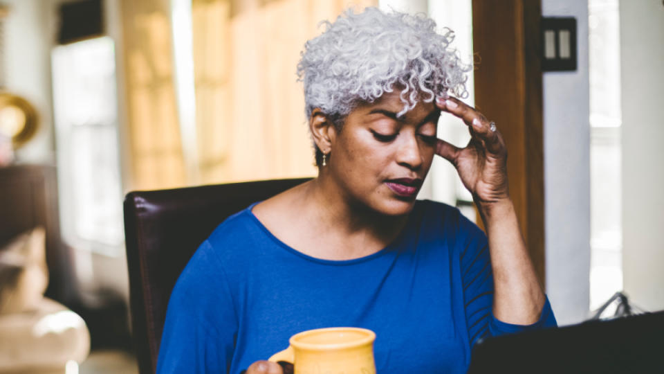 A woman with grey hair in a blue shirt holding her hand to her head due to stress