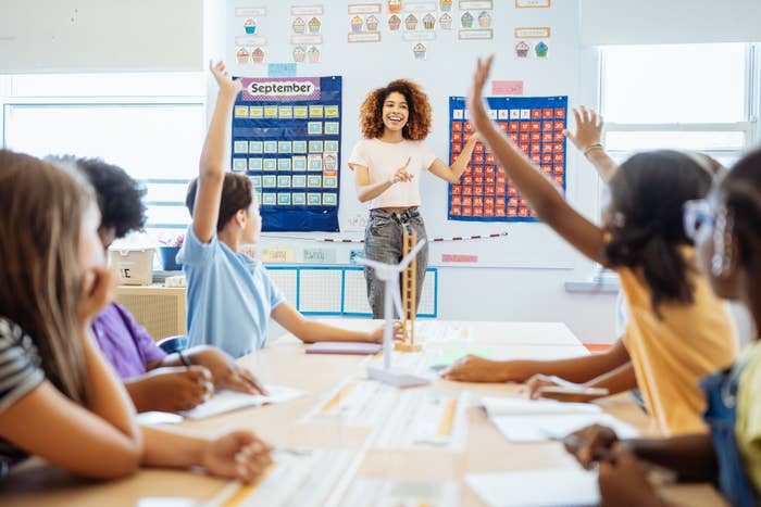 A teacher stands in front of a classroom with several students raising their hands. There are educational charts on the walls. The teacher smiles while pointing