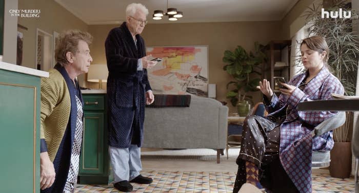 Steve Martin, Martin Short, and Selena Gomez in a living room, all dressed in robes. Selena is sitting and looking at her phone, while Steve and Martin are standing