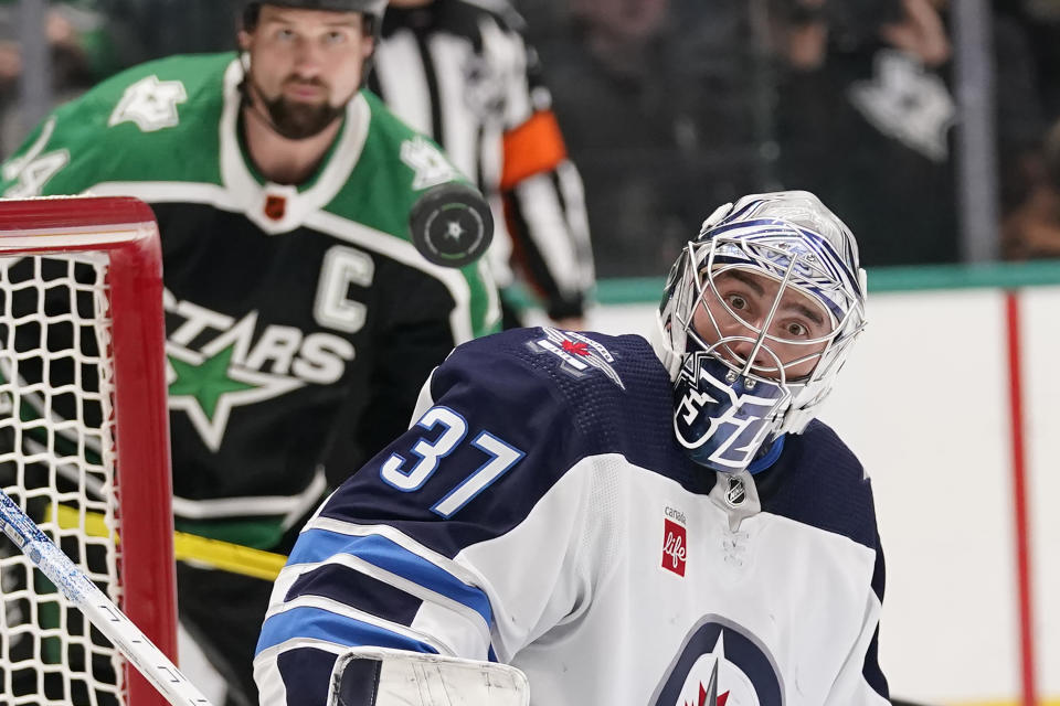 The puck defects off of Winnipeg Jets goaltender Connor Hellebuyck (37) as Dallas Stars left wing Jamie Benn (14) looks on during the first period of an NHL hockey game in Dallas, Friday, Nov. 25, 2022. (AP Photo/LM Otero)