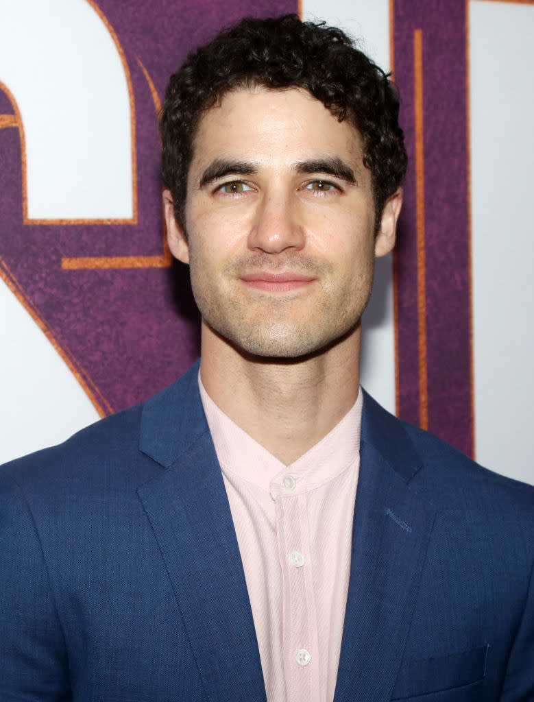 Darren Criss is wearing a blue suit over a light pink shirt, posing in front of a backdrop at a formal event