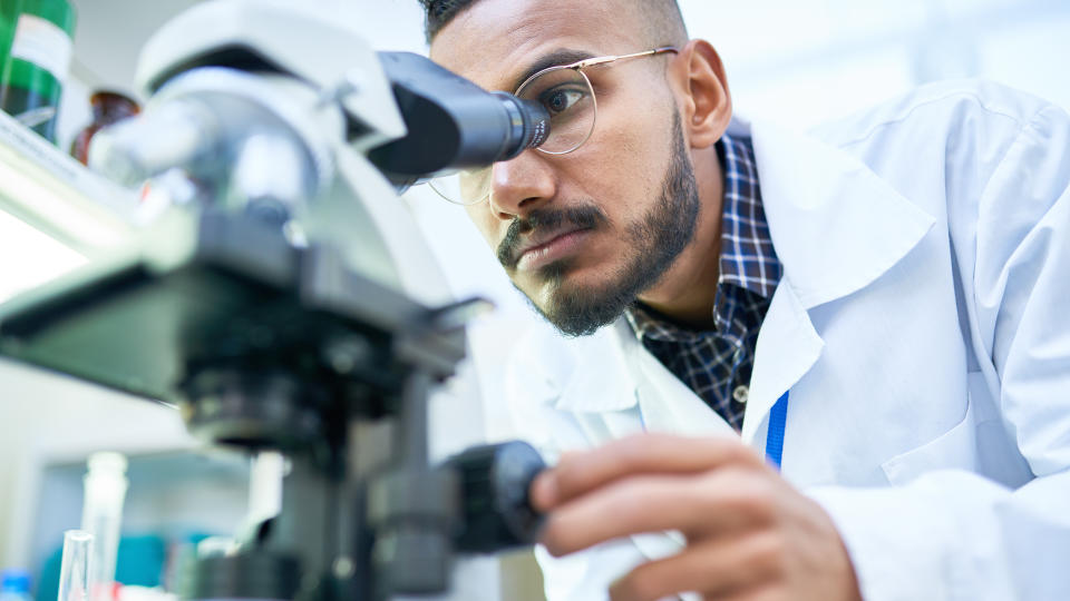Portrait of young Middle-Eastern scientist looking in microscope while working on medical research in science laboratory, copy space.